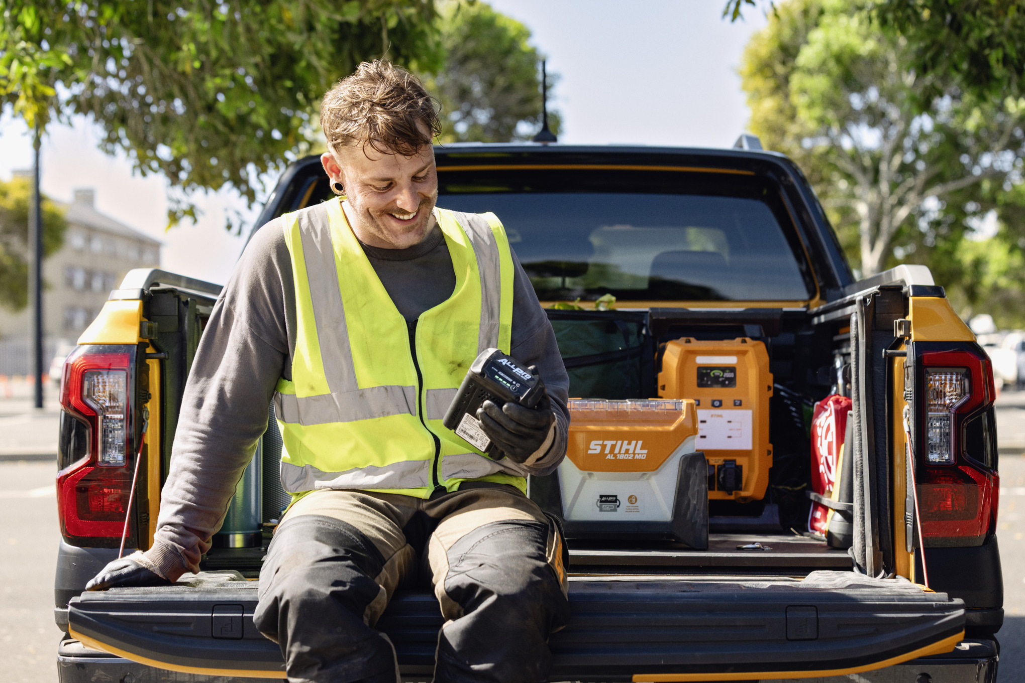 Man sitting on a pickup truck tailgate checking the charge level of his ALLPRO battery. In the background are the STIHL AL 1802 MO quick charger and the PS 3000 mobile power station