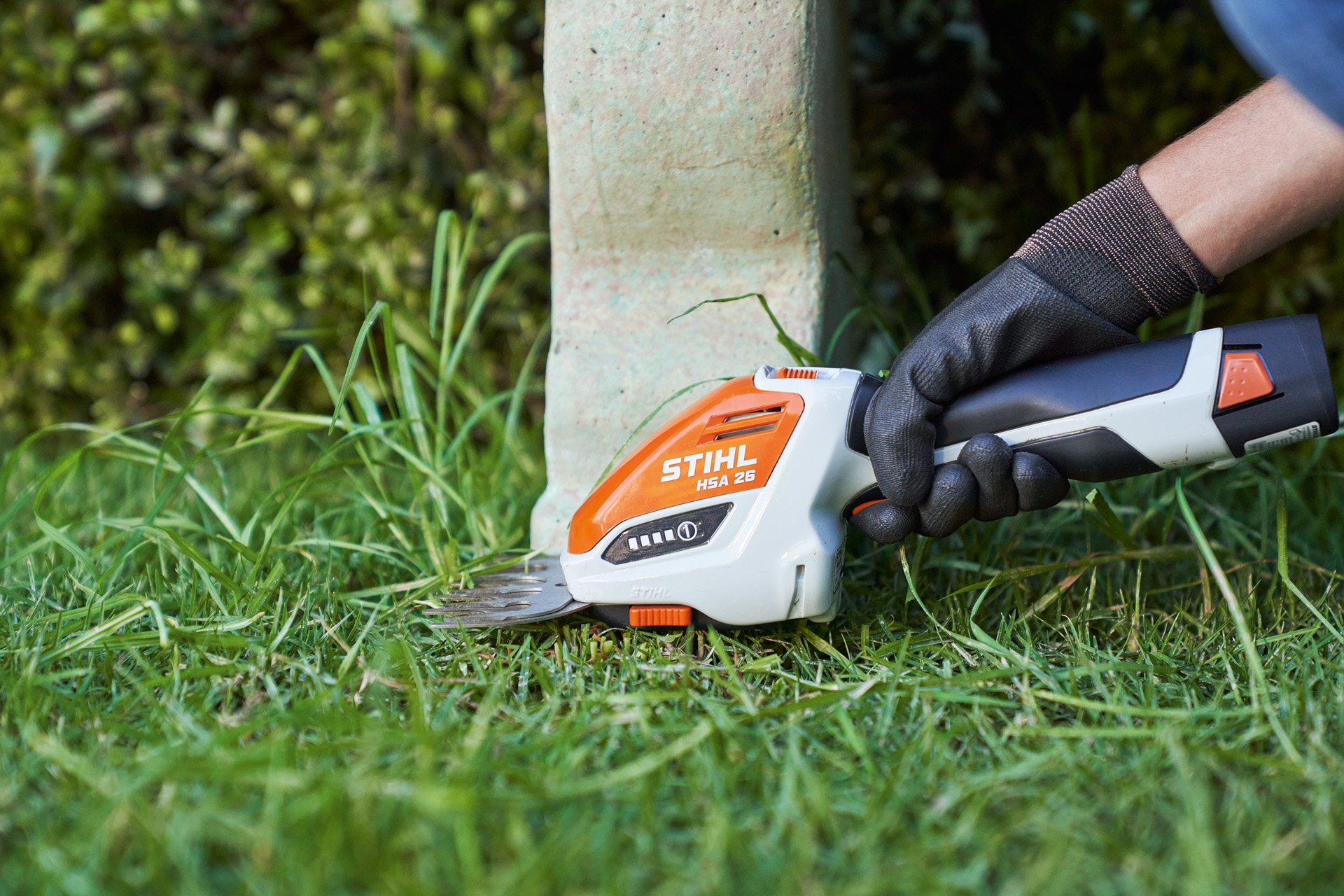 Close-up: hand wearing a protective glove trims lawn edges around a bench using the STIHL HSA 26 cordless shrub shears