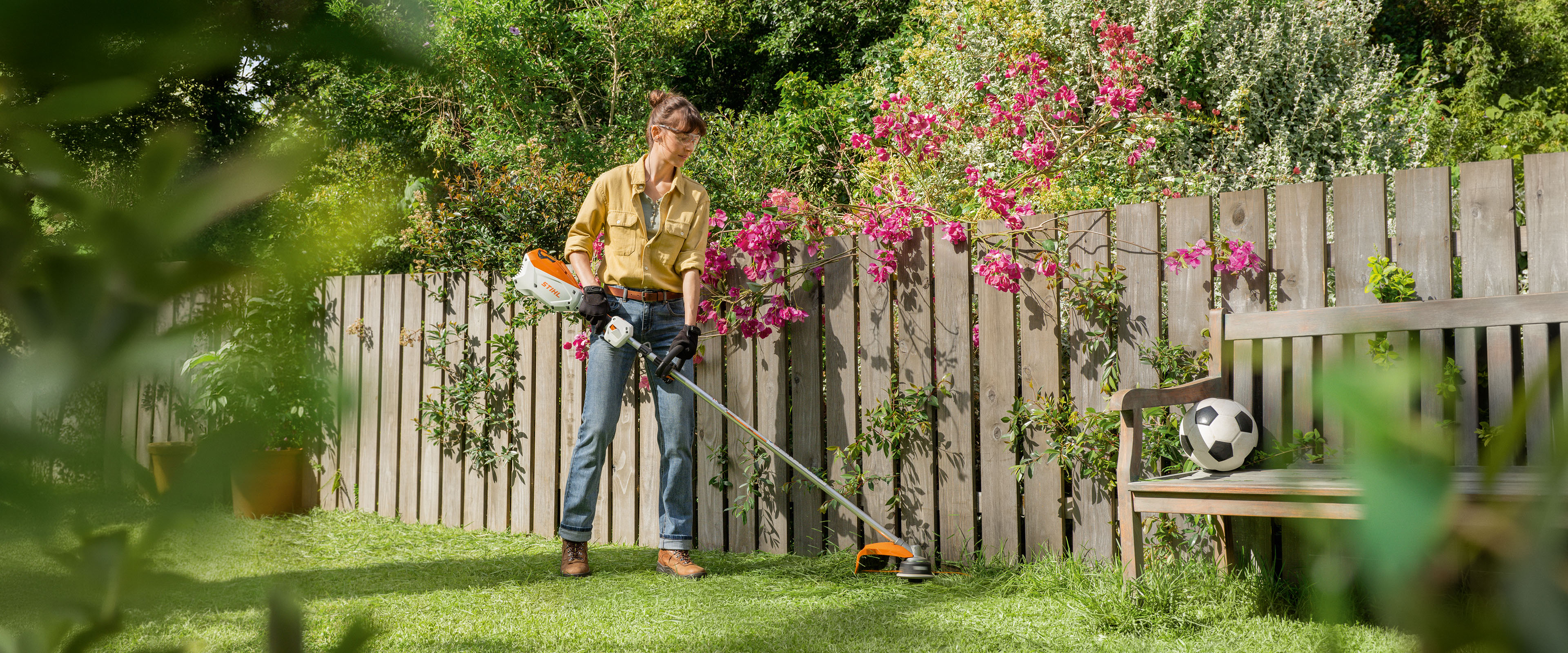Person using a STIHL string trimmer to cut grass along a backyard fence with plants and a bench nearby