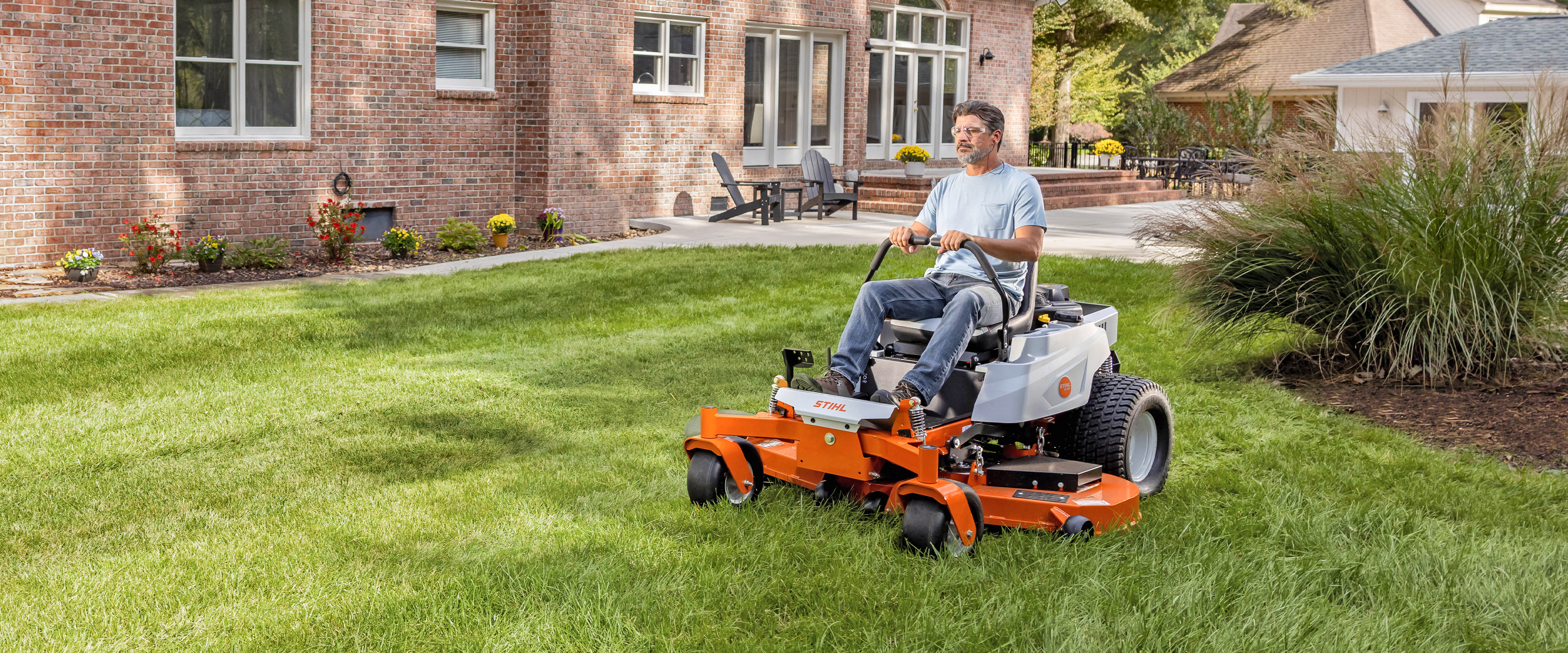 Homeowner riding a STIHL zero-turn mower across a lawn in front of a brick home.