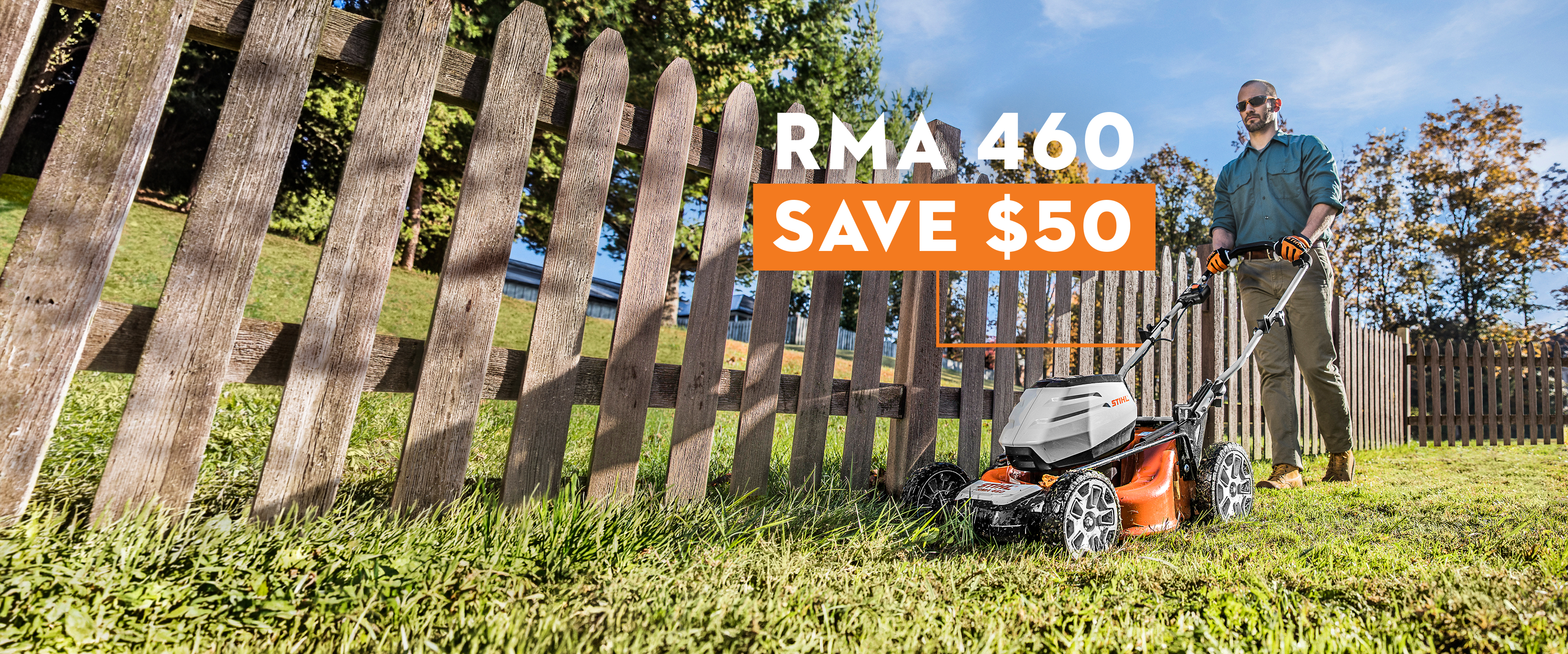 Person operating a self-propelled RMA 460 lawn mower across a neatly trimmed grassy yard alongside a tall wooden picket fence, with trees and a clear blue sky in a bright, sunny residential setting..