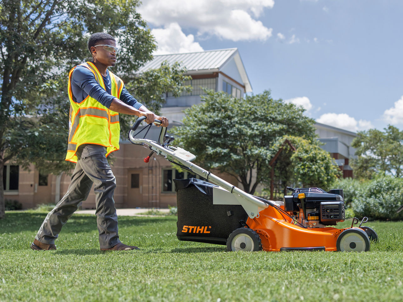 Landscape professional in a safety vest pushing a STIHL gas push mower with a rear grass bag.